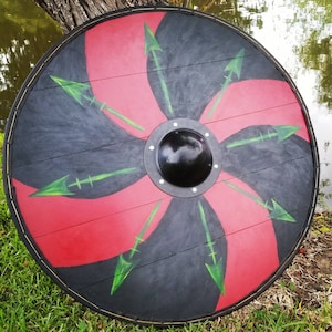 May include: A round wooden shield with a black rim and a central boss. The shield features a design of alternating black and red sections, with green arrows radiating from the center. The shield is suitable for display or costume.