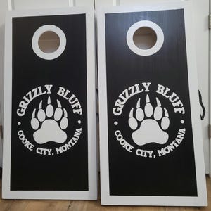 May include: Two rectangular cornhole boards with white frames and black playing surfaces. Each board features a circular hole at the top and a white graphic of a bear paw print surrounded by the text Grizzly Bluff Cooke City, Montana.