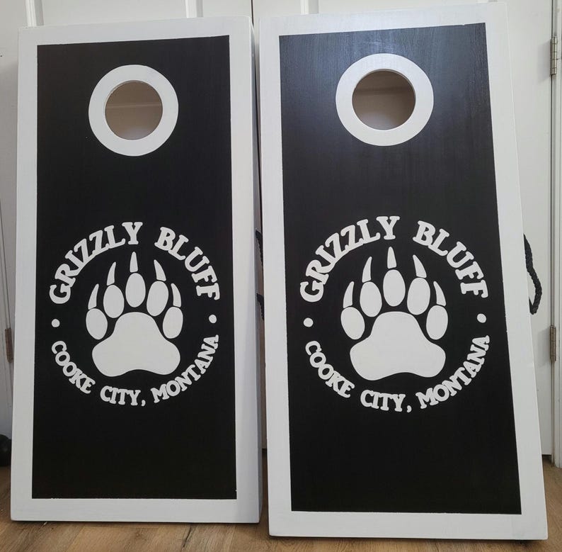 May include: Two rectangular cornhole boards with white frames and black playing surfaces. Each board features a circular hole at the top and a white graphic of a bear paw print surrounded by the text Grizzly Bluff Cooke City, Montana.