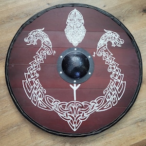 May include: Round wooden shield with a dark red stain and white Viking-style artwork. The shield features a central boss, a black rim, and intricate knotwork and dragon designs. A decorative piece for historical enthusiasts.