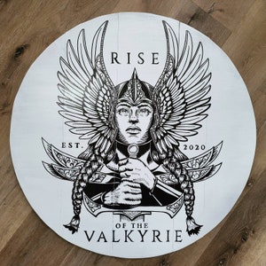 May include: A round, white wooden sign featuring a black and white illustration of a Valkyrie. The artwork includes the text "RISE OF THE VALKYRIE" and "EST. 2020". The Valkyrie is depicted with wings, a helmet, and a sword.