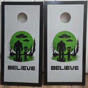 May include: A pair of rectangular cornhole boards with black frames and white surfaces. Each board features a circular hole at the top and a graphic of a Bigfoot and an alien standing before a large green moon with UFOs, trees, and the text BELIEVE.
