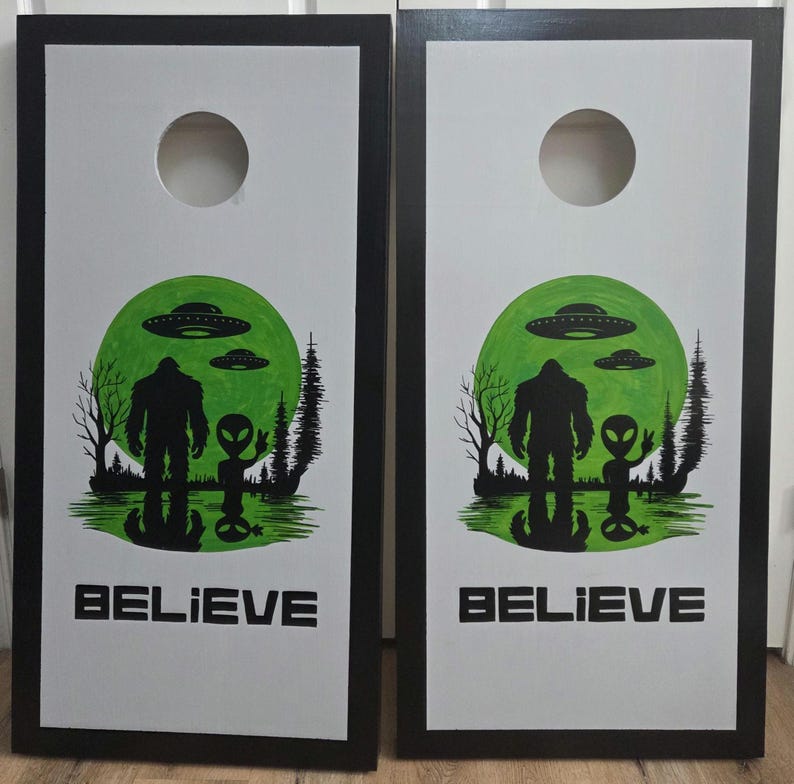 May include: A pair of rectangular cornhole boards with black frames and white surfaces. Each board features a circular hole at the top and a graphic of a Bigfoot and an alien standing before a large green moon with UFOs, trees, and the text BELIEVE.