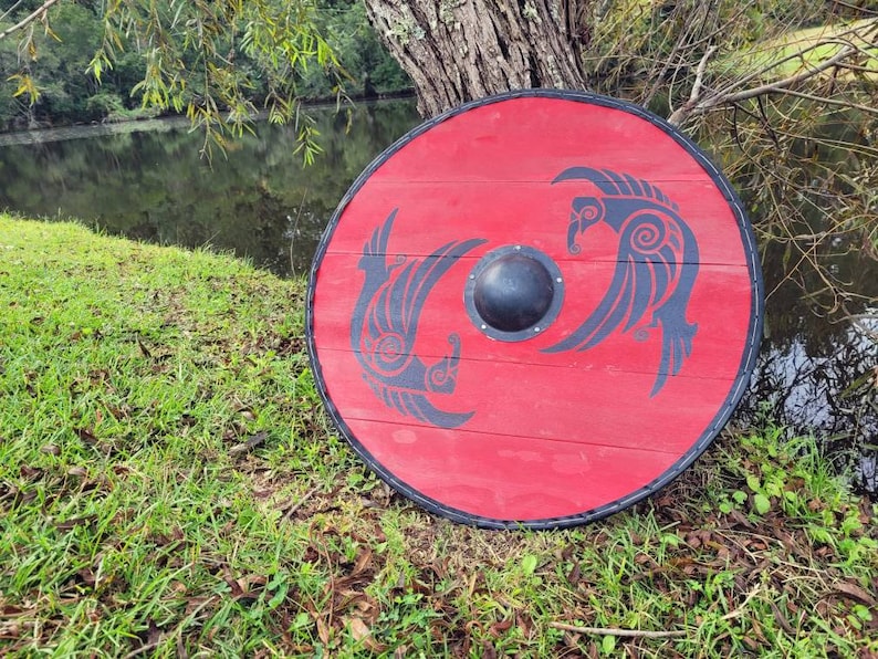 May include: A round, red wooden shield with a black rim and a central black boss. Two stylized black bird designs are painted on the shield. The shield is resting on green grass near a body of water.