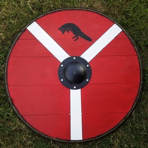 May include: A round, red wooden shield with a black fox silhouette jumping in the upper center. The shield features a white Y-shaped design and a black metal boss in the center, with a dark brown border.