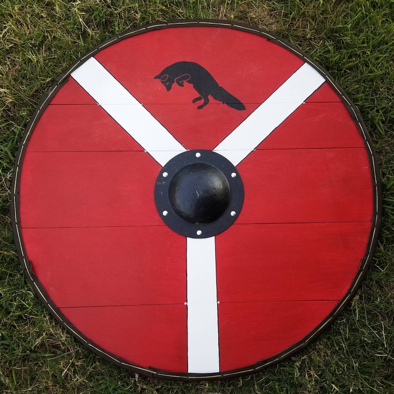 May include: A round, red wooden shield with a black fox silhouette jumping in the upper center. The shield features a white Y-shaped design and a black metal boss in the center, with a dark brown border.