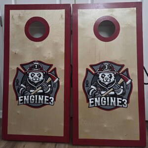 May include: Two rectangular wooden cornhole boards with a natural light wood finish and deep red painted borders. Each board features a circular hole at the top and a graphic of a panda wearing a firefighter helmet holding an axe with the text ENGINE3.