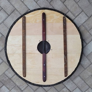 May include: A round wooden shield with a light wood grain center and a dark rim. The shield has three vertical wooden supports and two central, dark-stained wooden handles. A metal boss is in the center.