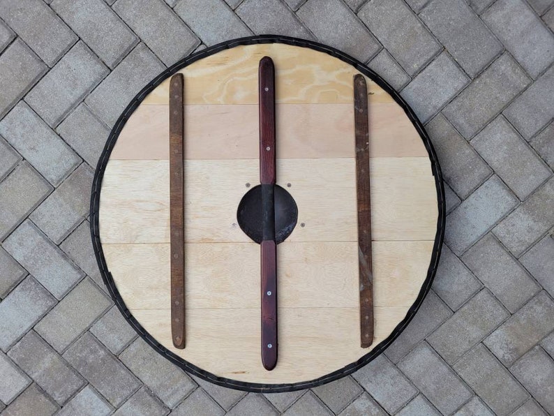 May include: A round wooden shield with a light wood grain center and a dark rim. The shield has three vertical wooden supports and two central, dark-stained wooden handles. A metal boss is in the center.