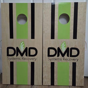 May include: Two light-coloured wooden cornhole boards featuring a central lime green stripe flanked by two thin black vertical stripes. Each board has a circular hole at the top and displays the black text DMD Systems Recovery with a green leaf icon.