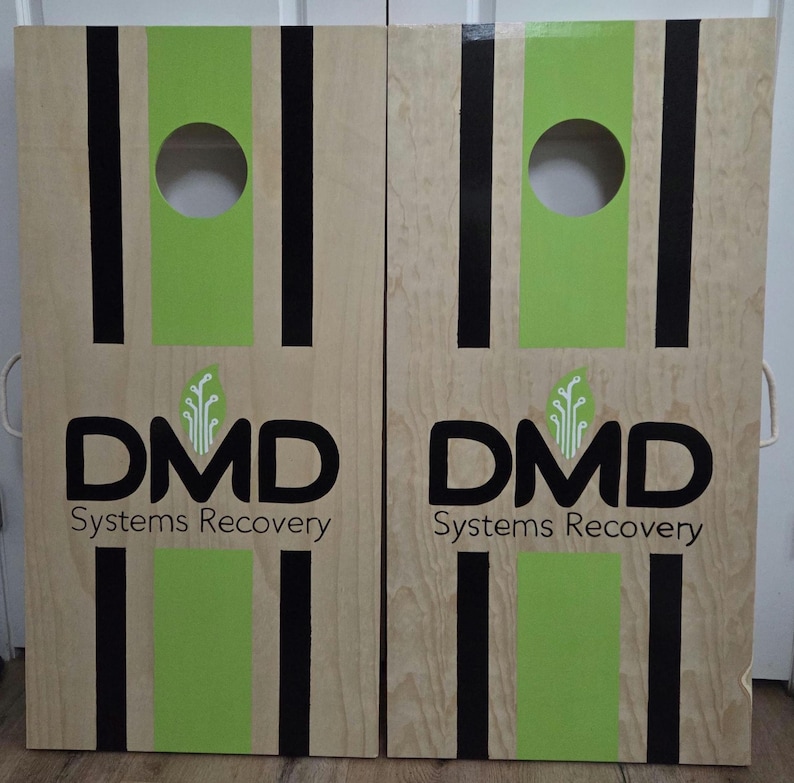 May include: Two light-coloured wooden cornhole boards featuring a central lime green stripe flanked by two thin black vertical stripes. Each board has a circular hole at the top and displays the black text DMD Systems Recovery with a green leaf icon.