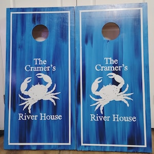 May include: Two rectangular cornhole boards with a blue and dark blue brushed finish. Each board features a circular hole at the top, a white border, and a white graphic of a crab. The text "The Cramer's River House" is printed in white.