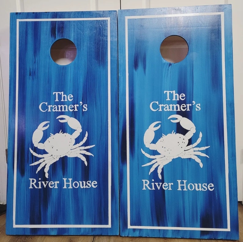May include: Two rectangular cornhole boards with a blue and dark blue brushed finish. Each board features a circular hole at the top, a white border, and a white graphic of a crab. The text "The Cramer's River House" is printed in white.