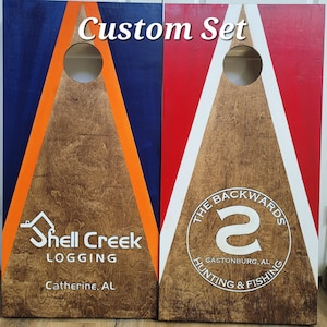 May include: Two rectangular wooden cornhole boards with circular holes at the top. The left board is blue with an orange triangle and the text Shell Creek Logging Catherine, AL. The right board is red with a white triangle and The Backwards S logo.