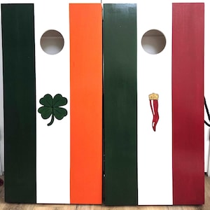 May include: Two rectangular wooden cornhole boards featuring vertical stripes. The left board has green, white, and orange stripes with a green four-leaf clover. The right board has green, white, and red stripes with a small red horn-shaped charm.