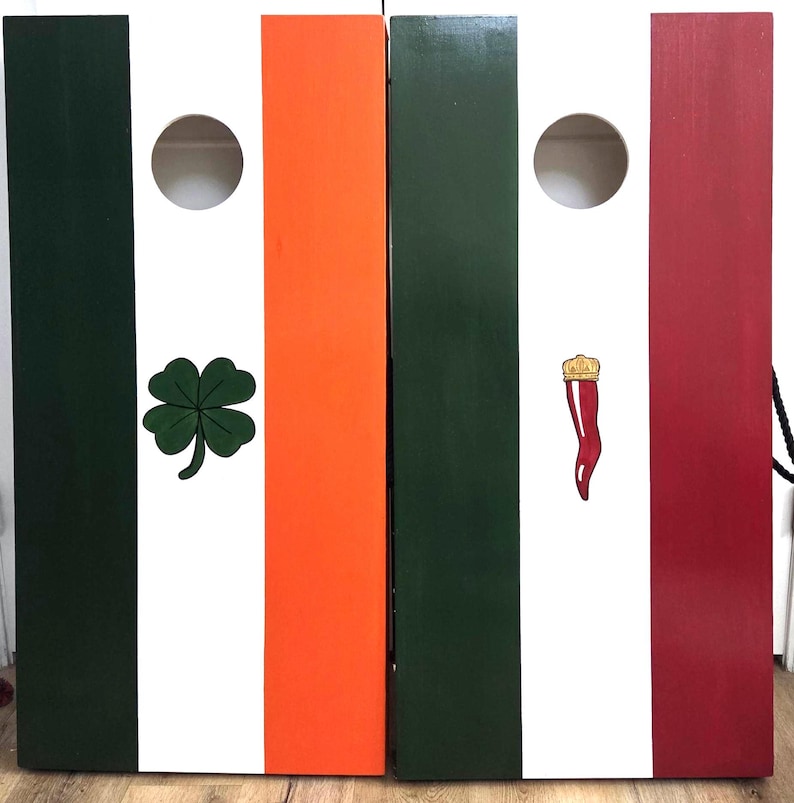 May include: Two rectangular wooden cornhole boards featuring vertical stripes. The left board has green, white, and orange stripes with a green four-leaf clover. The right board has green, white, and red stripes with a small red horn-shaped charm.