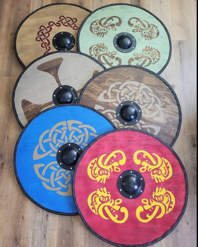 May include: A collection of six round wooden shields with black rims and central black bosses. Each shield features a different design in various colors, including red, blue, green, and brown, with Celtic knot and animal motifs.