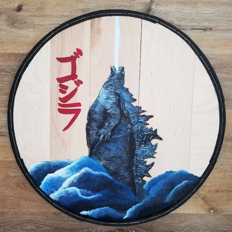 May include: Circular wooden art piece featuring a blue Godzilla illustration with a white beam emanating from its mouth. The Japanese word "Godzilla" is written in red. The artwork is framed with a black border.