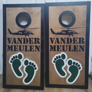 May include: A pair of rectangular wooden cornhole boards with black frames and natural wood surfaces. Each board features a black circular hole, a black silhouette of a helicopter, the text VANDER MEULEN, and two green footprints with white outlines.