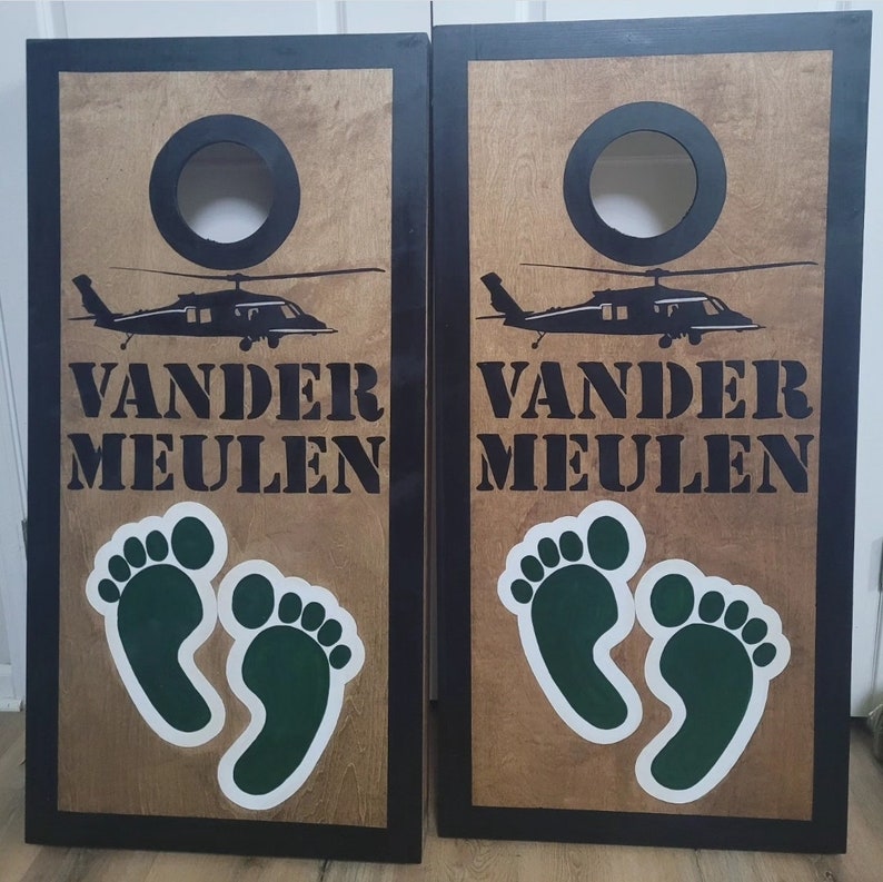 May include: A pair of rectangular wooden cornhole boards with black frames and natural wood surfaces. Each board features a black circular hole, a black silhouette of a helicopter, the text VANDER MEULEN, and two green footprints with white outlines.