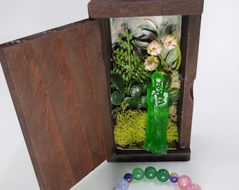 May include: A wooden box with a green figurine inside, surrounded by moss and greenery. The box is open, revealing the interior. A beaded bracelet with a silver charm hangs below the box.