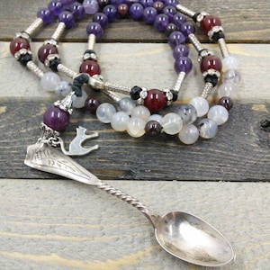 Puede incluir: Una cuchara de plata con mango retorcido y un dije de pájaro cuelga de un collar de cuentas. El collar está hecho de cuentas de amatista, cornalina y ágata blanca.