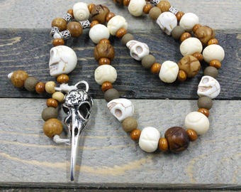 May include: A long necklace made of brown, white, and gray beads. The beads are shaped like skulls and have intricate carvings. A silver pendant shaped like a bird skull hangs from the necklace.