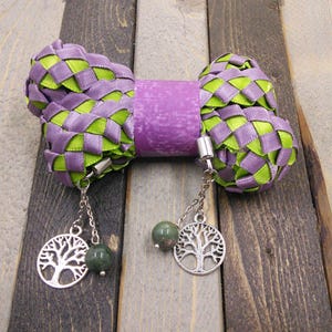 May include: A purple and green woven ribbon bow with two silver tree of life charms and green stone beads hanging from it.