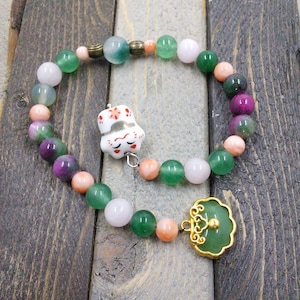 May include: A beaded bracelet featuring a variety of colorful beads in shades of green, pink, and purple. The bracelet includes a white ceramic cat charm and a green jade pendant with gold-colored accents. The bracelet is displayed on a wooden surface.