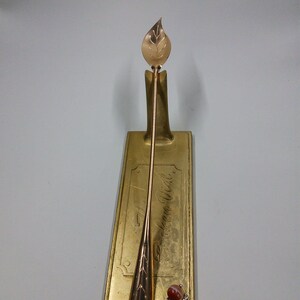 May include: A gold leaf-shaped spoon with a decorative design on the handle, displayed on a gold stand with the text "The Dream Wall". A small red gemstone charm hangs from the spoon.