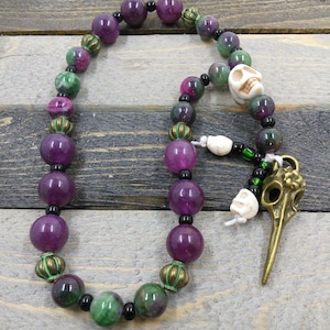 May include: A purple and green gemstone mala with a skull charm and a bird skull pendant. The mala is made with round beads and has a brass accent.