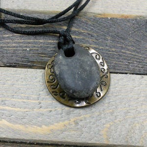May include: A black cord necklace with a round pendant. The pendant is made of two pieces: a dark gray stone and a gold-colored metal disc with a decorative pattern.