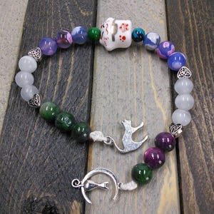 May include: A white ceramic charm with a cat design, surrounded by beads in shades of purple, green, and white. The charm is attached to a string of beads with a silver cat charm and a crescent moon charm.