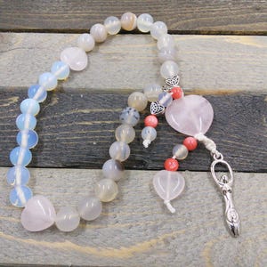 May include: A white and pink beaded mala necklace with a pink heart charm and a silver charm with a female figure. The necklace is on a wooden surface.
