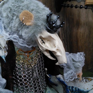 May include: A gray and white plush toy with a skull-like head, wearing a black crown and a silver chainmail necklace. The toy is dressed in a white lace collar and a green and blue patterned fabric.