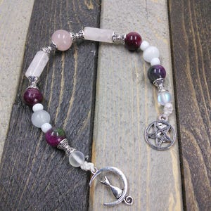 May include: A white and pink crystal beaded bracelet with a silver crescent moon charm and a pentagram charm. The bracelet is on a wooden surface.