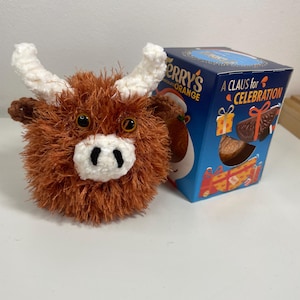 May include: A handcrafted, fluffy Highland cow toy with brown fur, white horns, and a white muzzle. Beside it is a blue box of Terry's Chocolate Orange, with the text "A CLAUS for CELEBRATION".