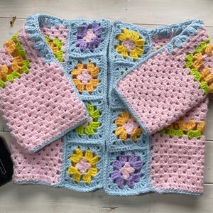 May include: A crochet cardigan constructed from light blue granny squares featuring floral motifs in yellow, pink, lavender, and lime green. The garment has long sleeves and a front-opening design, showcasing a multi-colored yarn pattern throughout.