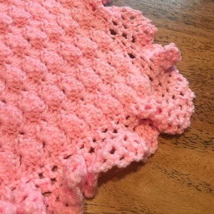 May include: A pink crocheted baby blanket with a scalloped edge. The blanket is made with a textured stitch and has a delicate, feminine look.