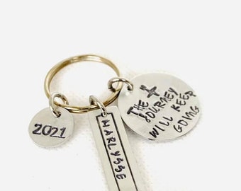 May include: A silver keychain with a gold ring holding three charms. One charm reads "2021", another says "The Journey Will Keep Going", and the third has the name "Marlysse". The charms are attached to a gold keyring.