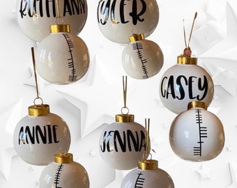 May include: Nine white Christmas baubles with gold tops and black lettering. The baubles are decorated with names, including Ruthann, Tyler, Casey, Annie, and Jenna. Some baubles have a black and white striped design.