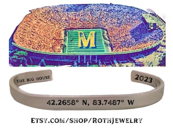 May include: A silver cuff bracelet with the text "THE BIG HOUSE" and "2023". The bracelet also features the coordinates 42.2658° N, 83.7487° W. The image includes a colorful illustration of a stadium and the text "MICHIGAN STADIUM COORDINATES".