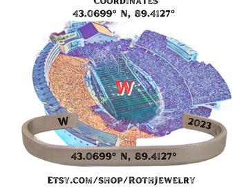 May include: A silver-colored cuff bracelet with the text "W", "2023", and coordinates 43.0699° N, 89.4127°. The bracelet is set against a backdrop of a stadium illustration. The text "Camp Randall Stadium Coordinates" is at the top.