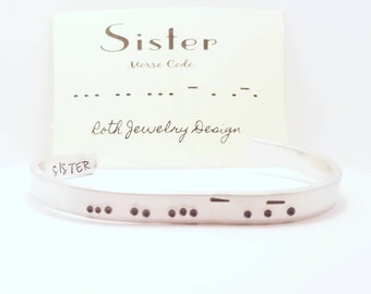 May include: A silver bangle bracelet with the word "Sister" engraved on it. The bracelet also features the word "Sister" in Morse code.