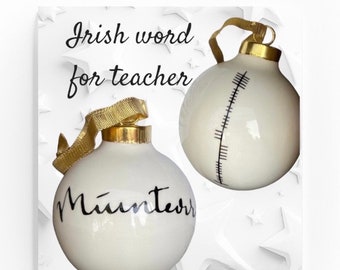 May include: Two white ceramic Christmas ornaments with gold ribbon. One ornament has the Irish word for teacher, "Múinteoir", written in black script. The other ornament has a black and white design of a ruler.