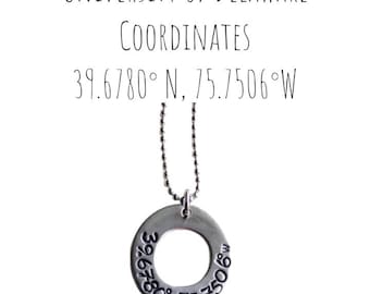 May include: A silver-colored pendant necklace with a circular charm. The charm has the coordinates 39.6780° N, 75.7506° W engraved around the edge. The text "UNIVERSITY OF DELAWARE COORDINATES" is above the pendant.