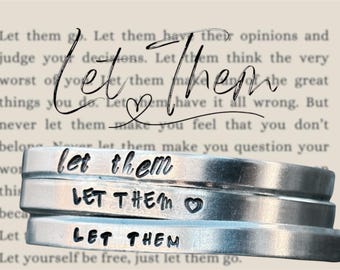 May include: Three silver metal bracelets stacked on top of each other, each stamped with the words "Let Them". The middle bracelet has a heart symbol. The background is a blurred image of text.