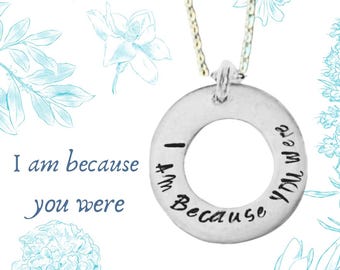May include: A silver necklace with a round pendant that says "I am because you were". The pendant is surrounded by a delicate chain. The necklace is on a white background with blue floral designs.