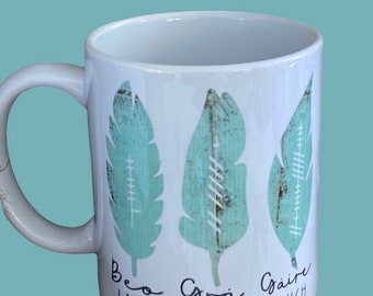 May include: White ceramic mug with three teal blue stylized leaves and the words "Beo Live", "Csrà Love", and "Gàire Laugh" printed on the bottom.
