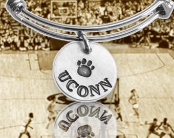 May include: A silver-colored bangle bracelet with two circular charms. Each charm has the word "UCONN" stamped on it, along with a paw print. The background is a sepia-toned image of a basketball game.